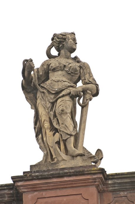 Die beige-braune Skulptur einer Frau in wallendem Gewand auf der Balustrade der Ludwigskirche. Ihr linker Arm stützt auf einen Anker, dessen Seil sie in beiden Händen hält.