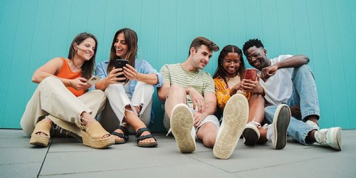 Schueler_Smartphones 5 Jugendliche mit 3 Smartphones sitzen an Wand gelehnt und schauen lächelnd auf ihre Smartphones