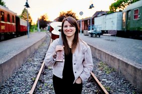 EASY – Isabell Krohn, Foto: Vincent Klein Eine junge Frau mit langem dunklem Haar steht bei fahlem Licht auf einem Bahngleis. Sie hält eine Gitarre, die auf ihrer Schulter ruht, und lächelt die Betrachtenden an.