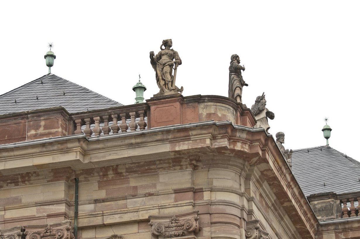 Blick von unten zum Dach der Ludwigskirche. Auf der Balustrade stehen mehrere beige-braune Steinskulpturen, darunter eine Frau in wallendem Gewand, die sich mit dem linken Arm auf einen Anker stützt.