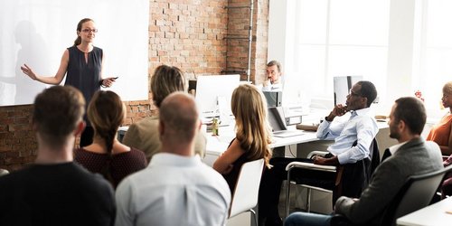 Mehrere Personen in einem Klassenraum hören der Konferenz Vorsitzenden zu Mehrere Personen in einem Klassenraum hören der Konferenz Vorsitzenden zu