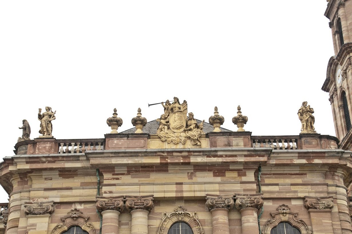 Blick von unten auf das Dach der Ludwigskirche. Auf der Balustrade stehen mehrere beige-braune Skulpturen. Links und rechts jeweils eine Frau in wallendem Gewand. Die Frau zur Linken hält Kreuz und Kelch, die Frau zur Rechten hält einen Säugling auf dem Arm und hat ein Kleinkind bei sich.