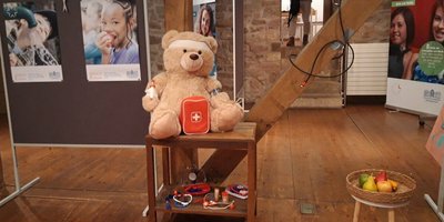 Ein Teddybär mit Verband und Spielzeug-Arztkoffer auf einem Hocker sitzend. Es handelt sich dabei um ein Exponat der Ausstellung zu Kinderrechten im Uhrmachershaus in Köllerbach.