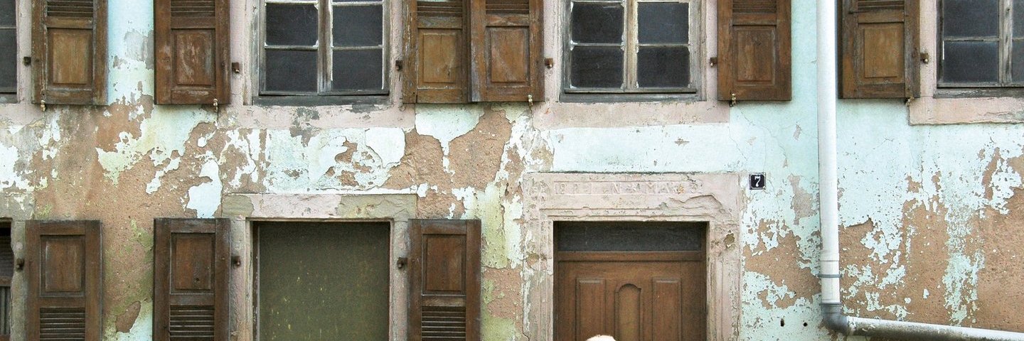 An einem alten Haus blättert die hellgrüne Wandfarbe ab. Die braunen, hölzernen Klappläden der vielen Fenster stehen offen.