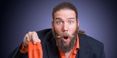 Ein Mann im Anzug, mit langen dunklen Bart und Haaren schaut überrascht in die Kamera. Sein linker Zeigefinger ist erhoben, in der rechten Hand hält er eine orangene Socke in die Luft.