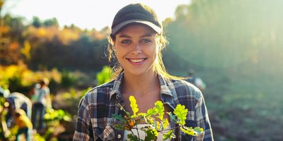 Frau mit Basecap lächelt in die Kamera. Im Hintergrund arbeiten Menschen auf einem Feld.