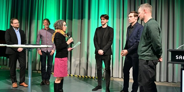 Die Jurymitglieder würdigen die Leistung und das Engagement von fugeefilms gGmbH. Foto: Stephan Hett/Regionalverband Saarbrücken Drei Jurymitglieder laudieren die Gründungsmitglieder von fugeefilms gGmbH auf der Bühne des Festsaals des Saarbrücker Schlosses.