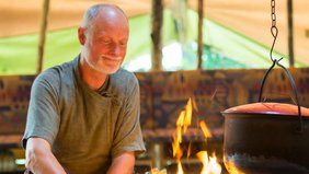 Ein Mann mit grauen Haaren und Halbglatze sitzt an einem Lagerfeuer in einem großen Zelt. Über dem Lagerfeuer hängt ein Topf. Der Mann legt in das Feuer blickend Holz nach.