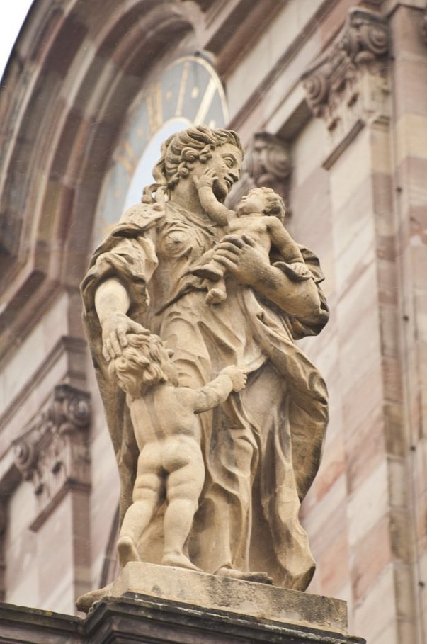 Die beige-braune Skulptur einer Frau in wallendem Gewand. Sie hält einen Säugling auf dem Arm und berührt mit der rechten Hand den Kopf eines nackten Kleinkindes, das nach ihrem Gewand greift.