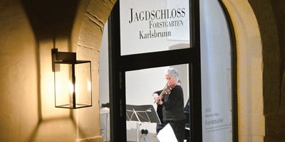 Blick durch ein Fenster in die Remise des Jagdschlosses, in der eine grauhaarige Frau Geige spielt.