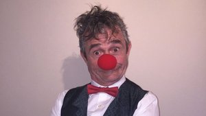 Ein Mann mit grauen kurzen Haaren trägt eine rote Clownsnase, ein weißes Hemd mit einer schwarzen Weste darüber. Er hat die Hände vor sich verschränkt und lacht in die Kamera.