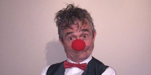 Ein Mann mit grauen kurzen Haaren trägt eine rote Clownsnase, ein weißes Hemd mit einer schwarzen Weste darüber. Er hat die Hände vor sich verschränkt und lacht in die Kamera.