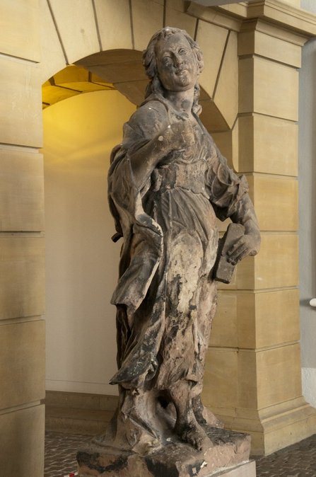 Die beige-braune Skulptur einer Frau in wallendem Gewand im Eingangsbereich des Saarbrücker Schlosses. In ihrer linken Hand hält sie die Überreste eines großen Kreuzes.