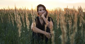 Eine Frau mit langen braunen Haaren und schwarzer Kleidung sitzt in einem Feld. Im Hintergrund ist der Sonnenuntergang zu sehen. Sie hat den Kopf nachdenklich auf eine Hand gestützt und schaut in die Ferne.