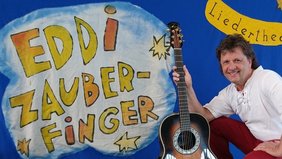 Ein Mann mit schulterlangen dunklen Haaren, weißem Hemd und roter Hose sitzt vor einem blauen Hintergrund auf dem Eddi Zauberfinger aufgemalt wurde. Er lacht in die Kamera und hält eine braune Akkustik-Gitarre in der Hand.
