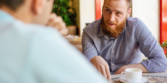 Blick über die Schuler eines Mannes, der am Tisch sitzt und mit einem jungen Mann ein Gespräch führt