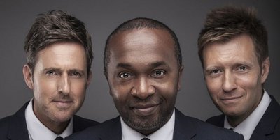 Drei Männer in schwarzen Anzügen schauen lachend in die Kamera. Die beiden äußeren tragen dunkleblondes, kurzes Haar. Der Mann in der Mitte trägt schwarzes sehr kurzes Haar.