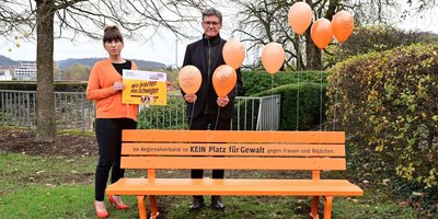 Orangene Bank im Schlossgarten mit dem Schriftzug "Im Regionalverband ist kein Platz für Gewalt gegen Frauen und Mädchen". Dahinter stehend Regionalverbandsdirektor Peter Gillo und die Frauenbeauftragte Mirjam Altmeier-Koletzki.