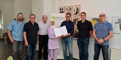 Regionalverbandsdirektor Peter Gillo überreicht an mehrere Mitglieder des Kulturvereins Burbach ein Förderschreiben für das Projekt "Hier daheim"
