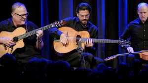 Drei Männer in schwarzen Hemden sitzen Gitarre spielend nebeneinander auf einer Bühne.