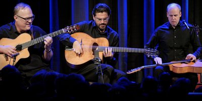 Drei Männer in schwarzen Hemden sitzen Gitarre spielend nebeneinander auf einer Bühne.