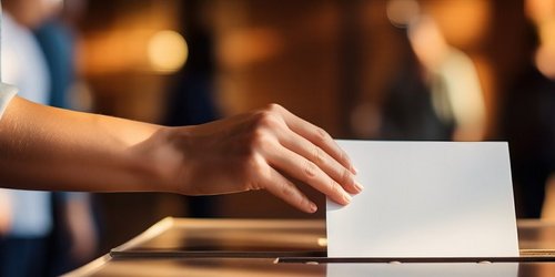 Hand einer jungen Frau die einen Stimmzettel in eine Wahlurne wirft. 