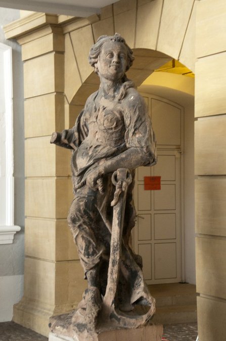 Die beige-braune Skulptur einer Frau in wallendem Gewand im Eingangsbereich des Saarbrücker Schlosses. Ihr linker Arm stützt auf einen Anker, dessen Seil sie in der linken Hand hält. Die Hand an ihrem rechten Arm fehlt.