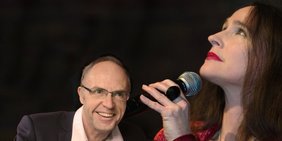 Vor schwarzem Hintergrund: Ein Mann in Anzug mit Brille und Halbglatze spielt lächelnd Klaiver. Rechts neben ihm hält eine Frau in dunkelrotem Samtkleid mit langem dunklem Haar und andächtigem Blick ein Mikrofon.