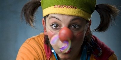 Eine Frau mit grüner Mütze und zwei Zöpfen schaut überrascht in die Kamera. Sie trägt eine rote Clownsnase, ein orang-blaues T-Shirt und hat den rechten Zeigefinger gehoben. Aus ihrem Mund kommt eine bunte Blase. 