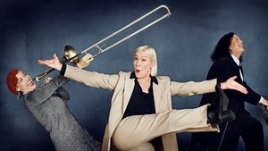 Drei Frauen stehen nebeneinander. Die linke Frau trägt eine rote Mütze, ein graues Kleid und hält eine Trompete vor sich ausggestreckt. Die mittlere blonde Frau trägt einen begen Anzug und hat beide Arme von sich gestreckt. Die rechte dunklehaarige Frau trägt einen schwarzen Anzug und sitzt lachend an einem Klavier.